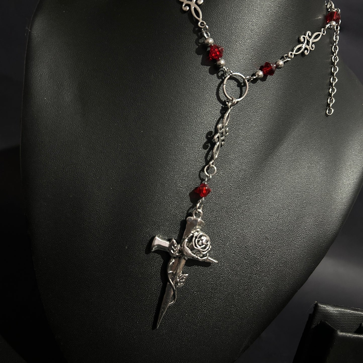 Close-up of silver gothic cross pendant wrapped in a rose, hanging from a Y-drop chain with red crystal beads and ornate filigree. A romantic gothic necklace perfect for alt, cosplay, and vampire-inspired looks.