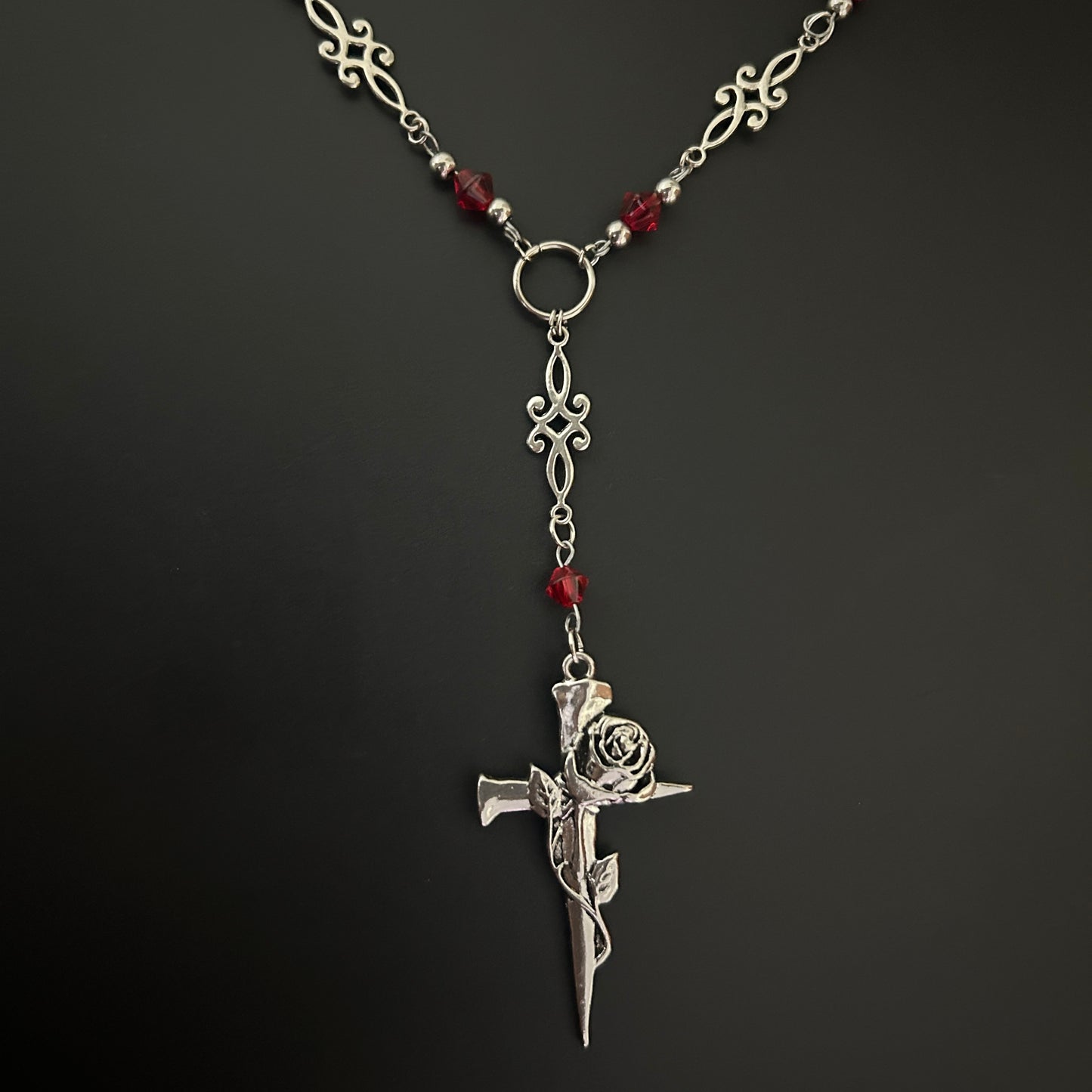 Flat-lay shot of gothic Y-drop necklace with ornate metalwork, red crystal beads, and a spiked rose-wrapped cross pendant. Gothic accessory suited for cosplay, alt fashion, and K-pop inspired outfits.
