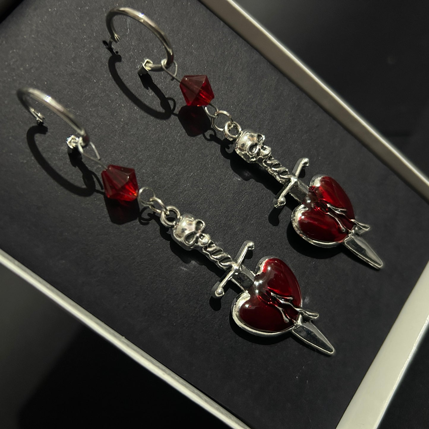 Skull Dagger Heart Earrings on silver hoops displayed in a box - close-up. Gothic style chrome plated stainless steel earrings for lovers of alt and punk fashion