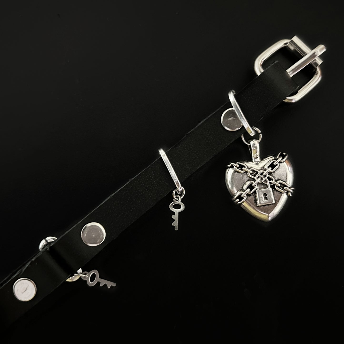 Flat lay of black leather choker with chained silver heart and padlock centerpiece, with stainless steel key charms. Gothic, alt, rockstar, and grunge jewelry by Phantom Cat India.