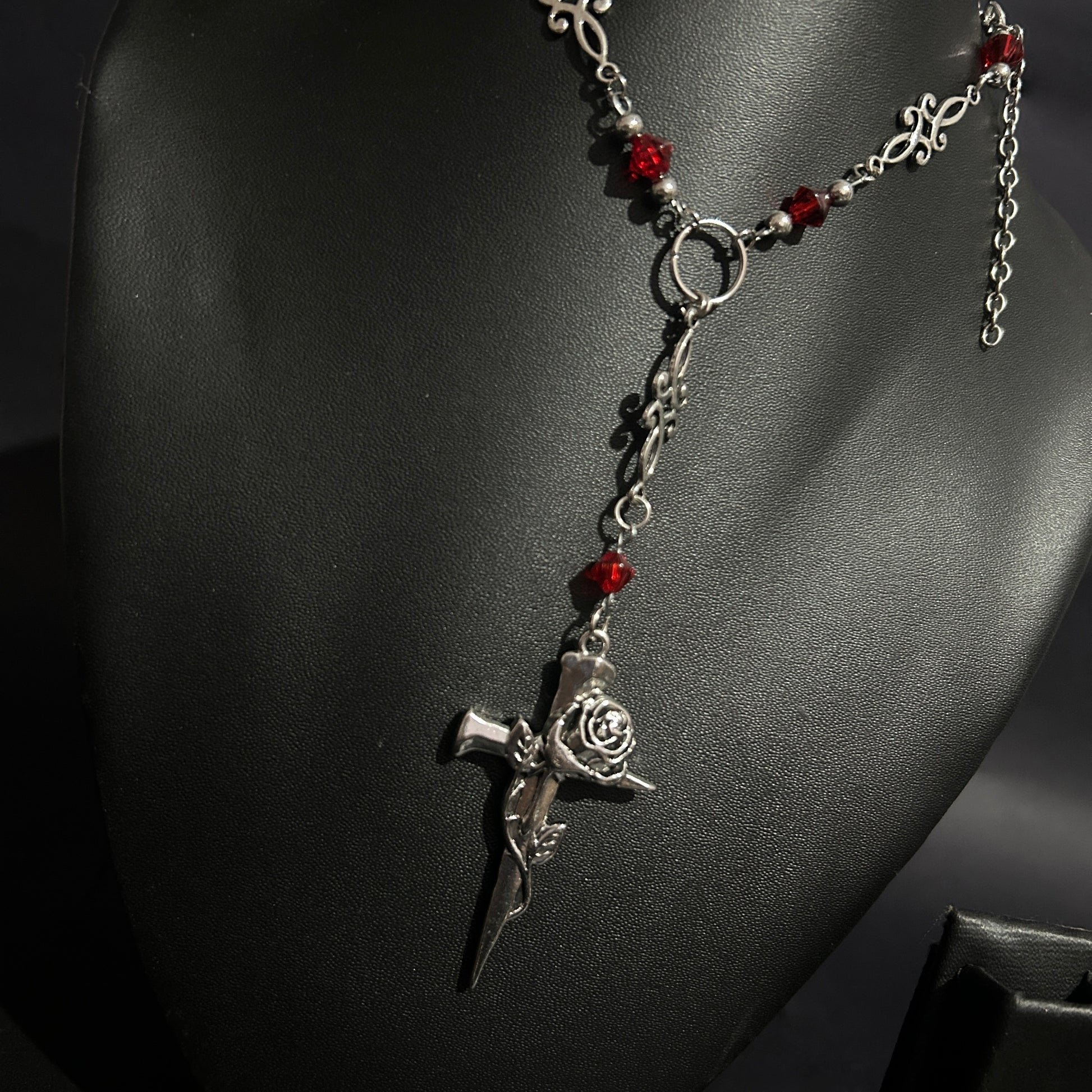 Close-up of silver gothic cross pendant wrapped in a rose, hanging from a Y-drop chain with red crystal beads and ornate filigree. A romantic gothic necklace perfect for alt, cosplay, and vampire-inspired looks.
