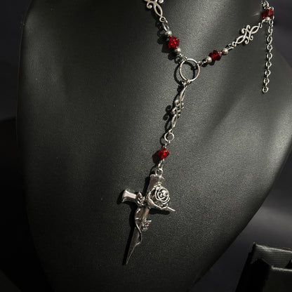 Close-up of silver gothic cross pendant wrapped in a rose, hanging from a Y-drop chain with red crystal beads and ornate filigree. A romantic gothic necklace perfect for alt, cosplay, and vampire-inspired looks.