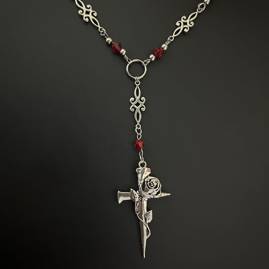 Flat-lay shot of gothic Y-drop necklace with ornate metalwork, red crystal beads, and a spiked rose-wrapped cross pendant. Gothic accessory suited for cosplay, alt fashion, and K-pop inspired outfits.