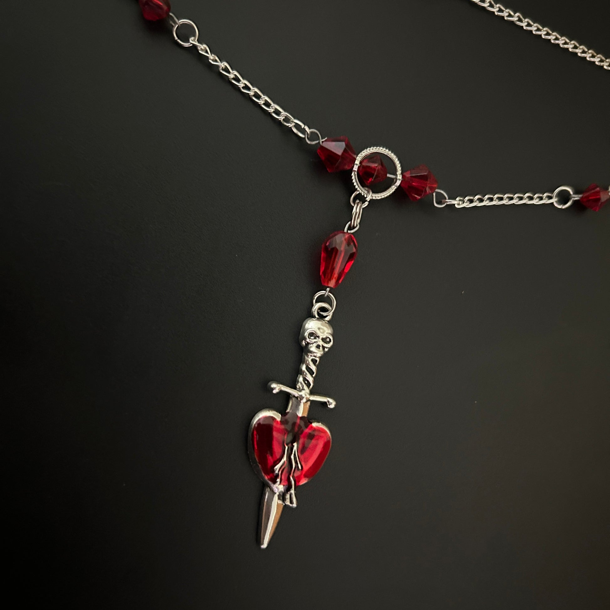 Flatlay of Crimson Heart Dagger Necklace with deep red stones and spear pendant. Gothic romance meets alt fashion jewelry.