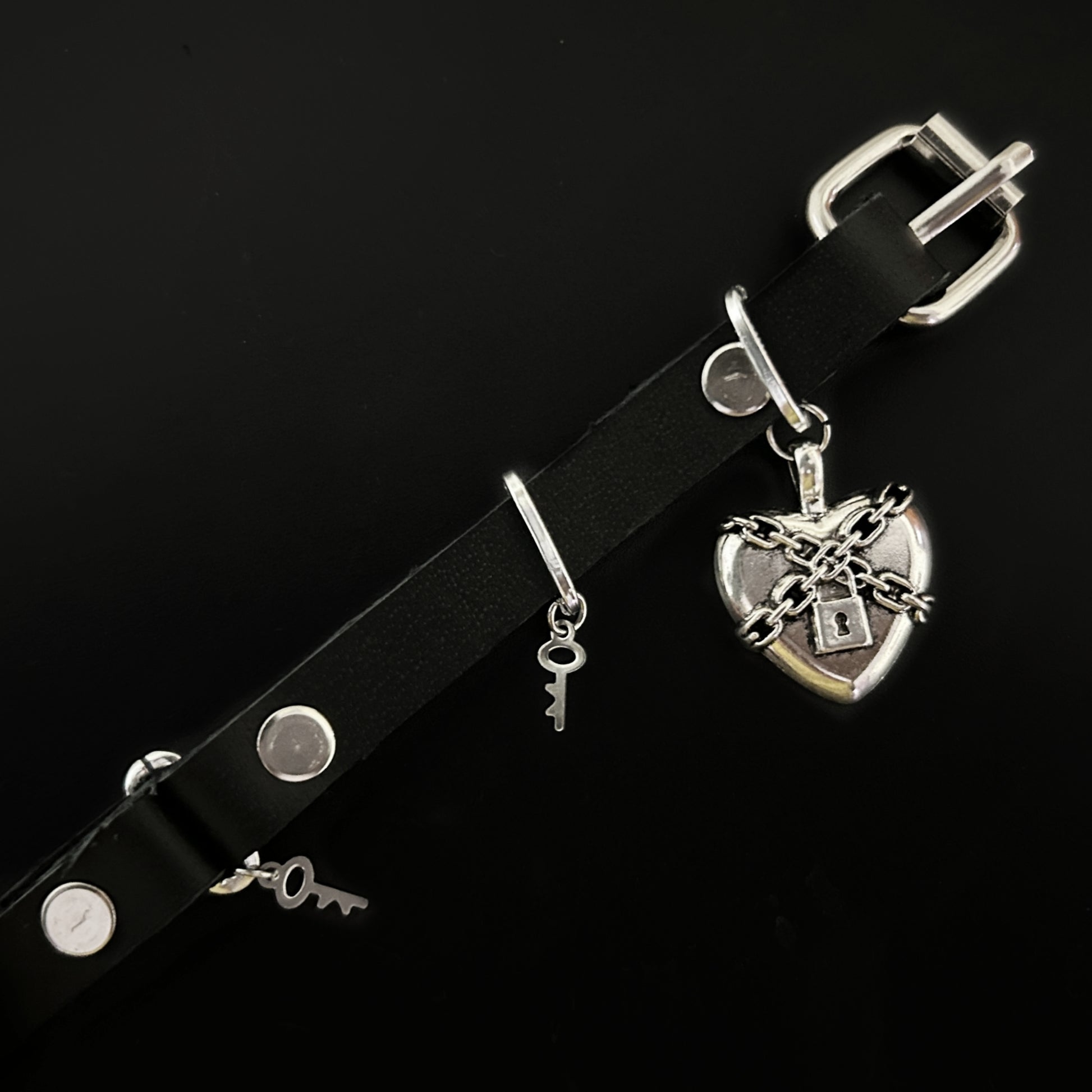 Flat lay of black leather choker with chained silver heart and padlock centerpiece, with stainless steel key charms. Gothic, alt, rockstar, and grunge jewelry by Phantom Cat India.