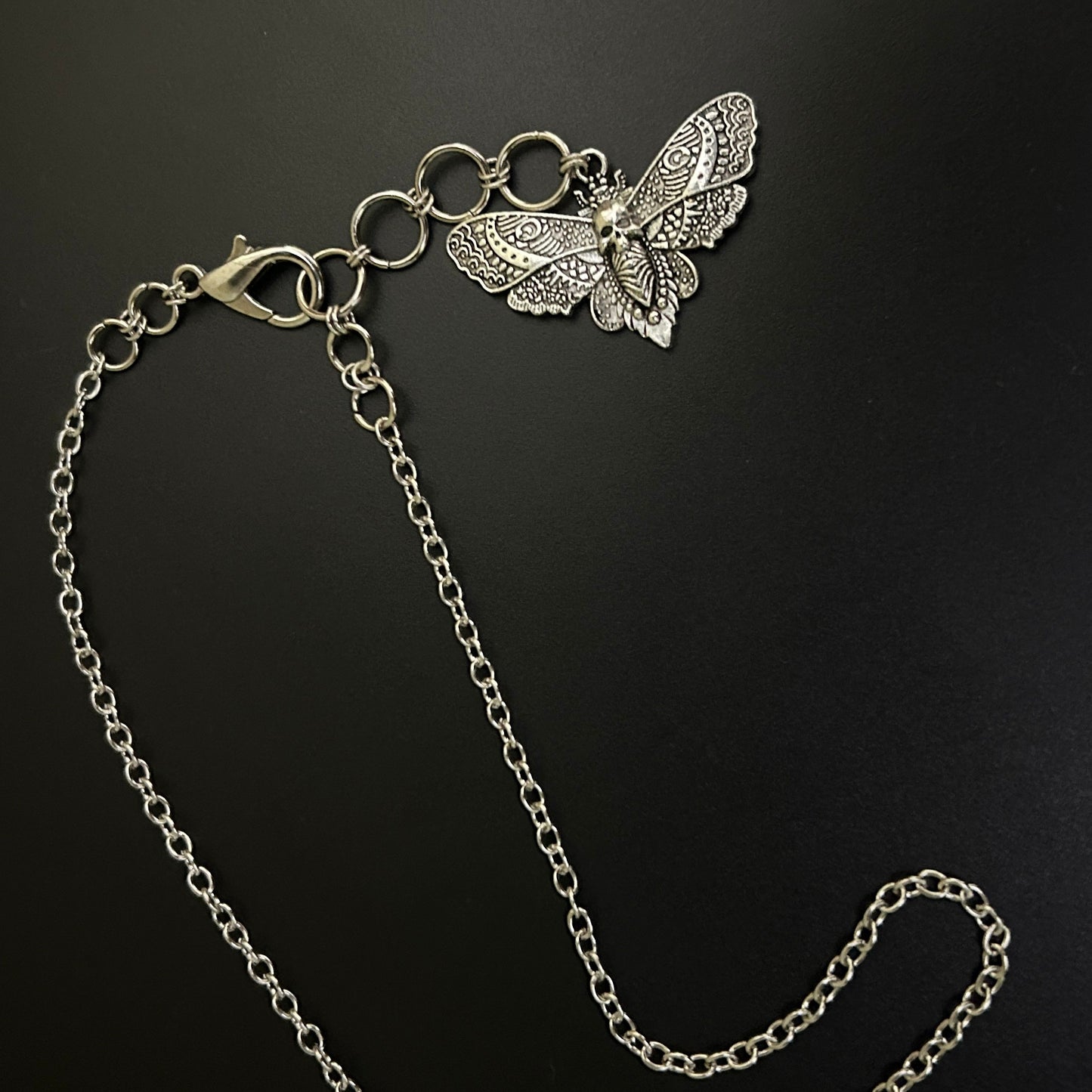 Flat lay of Shadowed Wings Skull Choker Necklace with silver chain and a drop extension holding a skull-winged moth charm. A gothic necklace ideal for alt fashion and grunge jewelry looks.