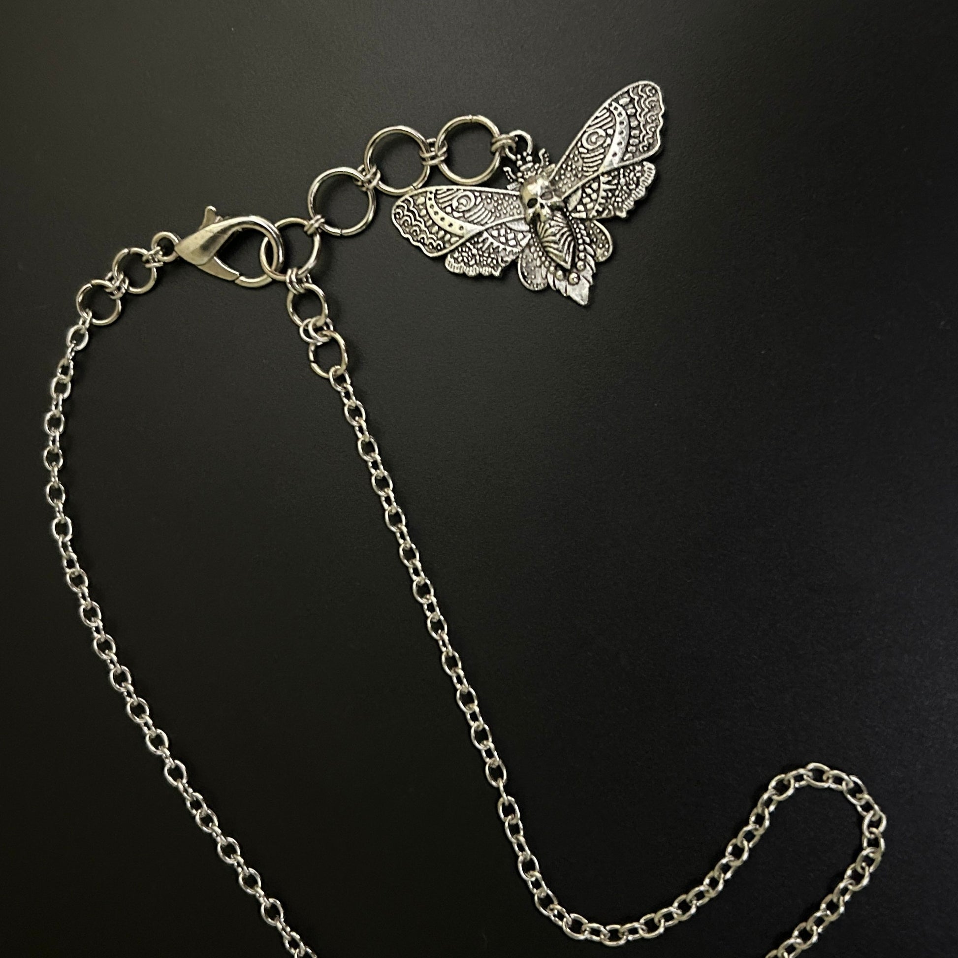 Flat lay of Shadowed Wings Skull Choker Necklace with silver chain and a drop extension holding a skull-winged moth charm. A gothic necklace ideal for alt fashion and grunge jewelry looks.
