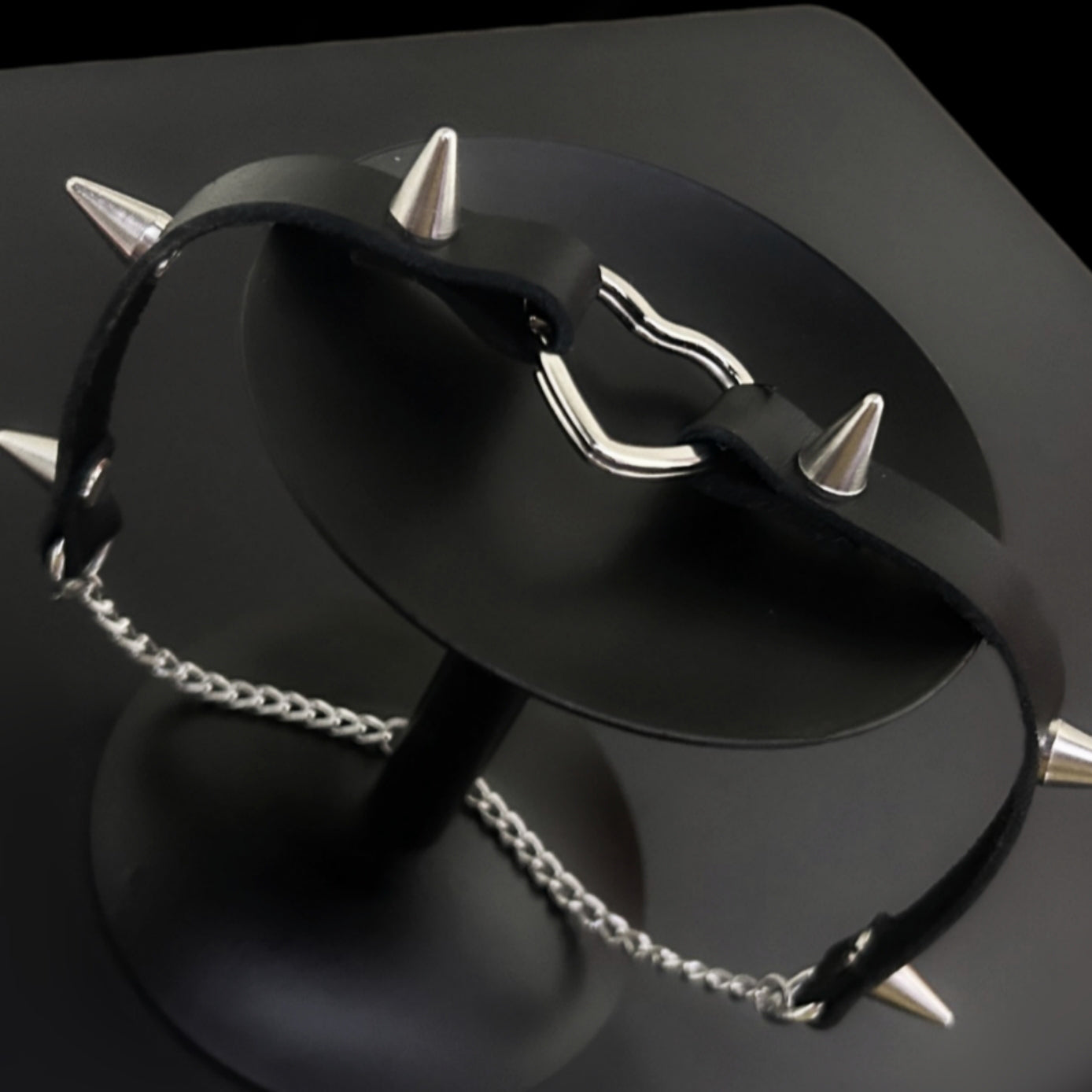 Flat lay of spiked leather choker with stainless steel heart, displayed on a black circular stand. Conical metal spikes. Ideal for gothic, punk, grunge, and alt fashion styling in India