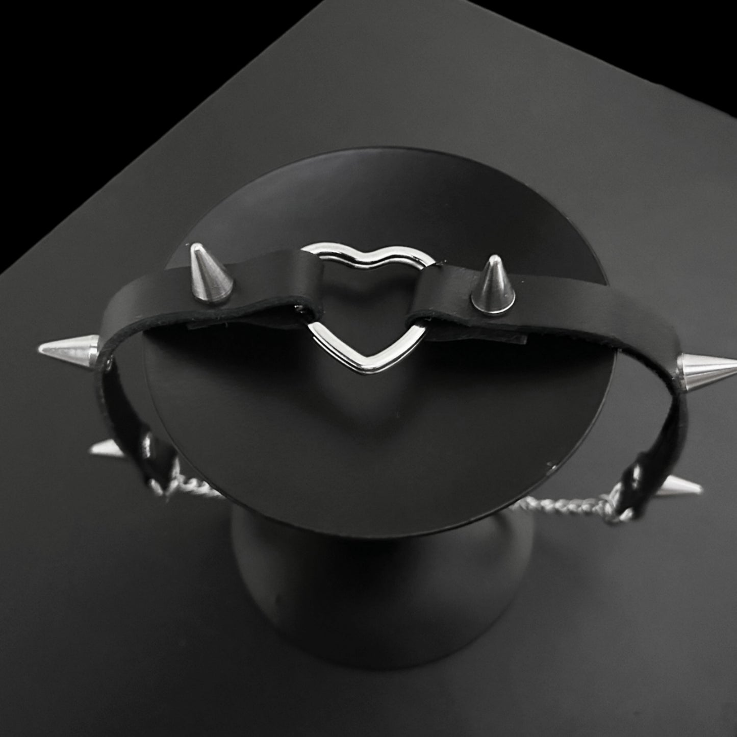 Top view flat lay of black leather gothic choker with bold conical spikes and a silver heart ring centerpiece, perfect for grunge jewelry lovers, Y2K styling, alt fashion, and cosplay outfits.