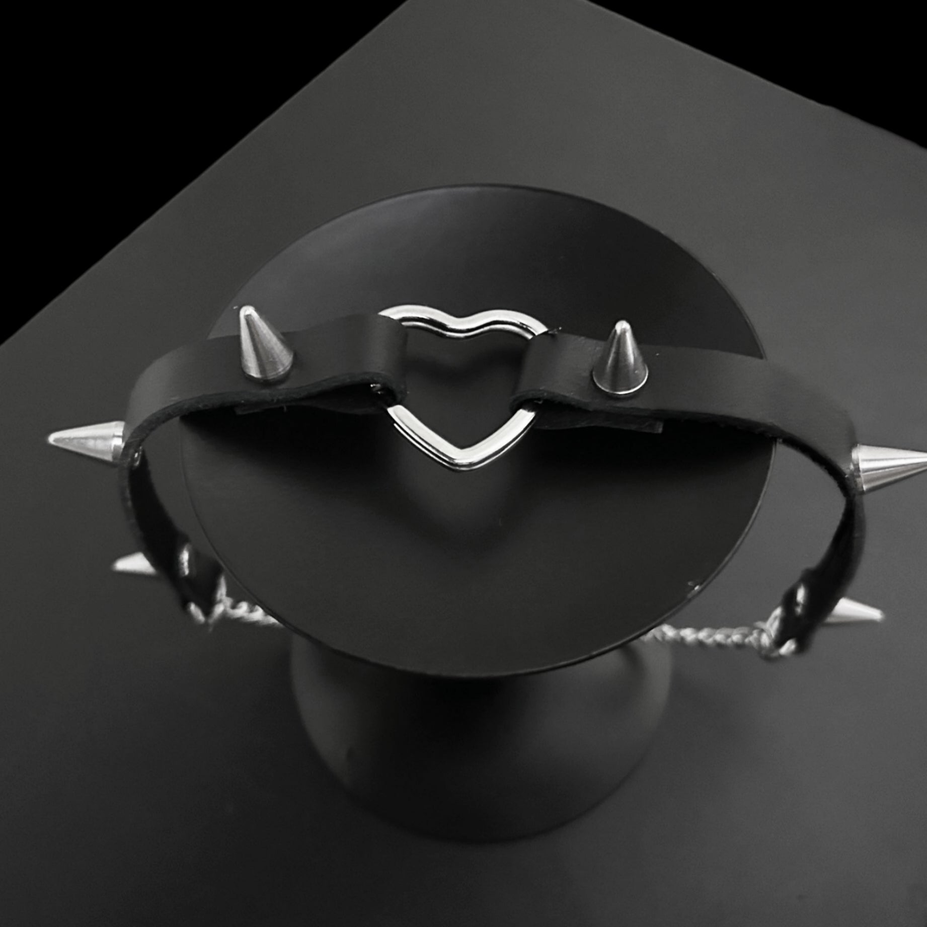 Top view flat lay of black leather gothic choker with bold conical spikes and a silver heart ring centerpiece, perfect for grunge jewelry lovers, Y2K styling, alt fashion, and cosplay outfits.