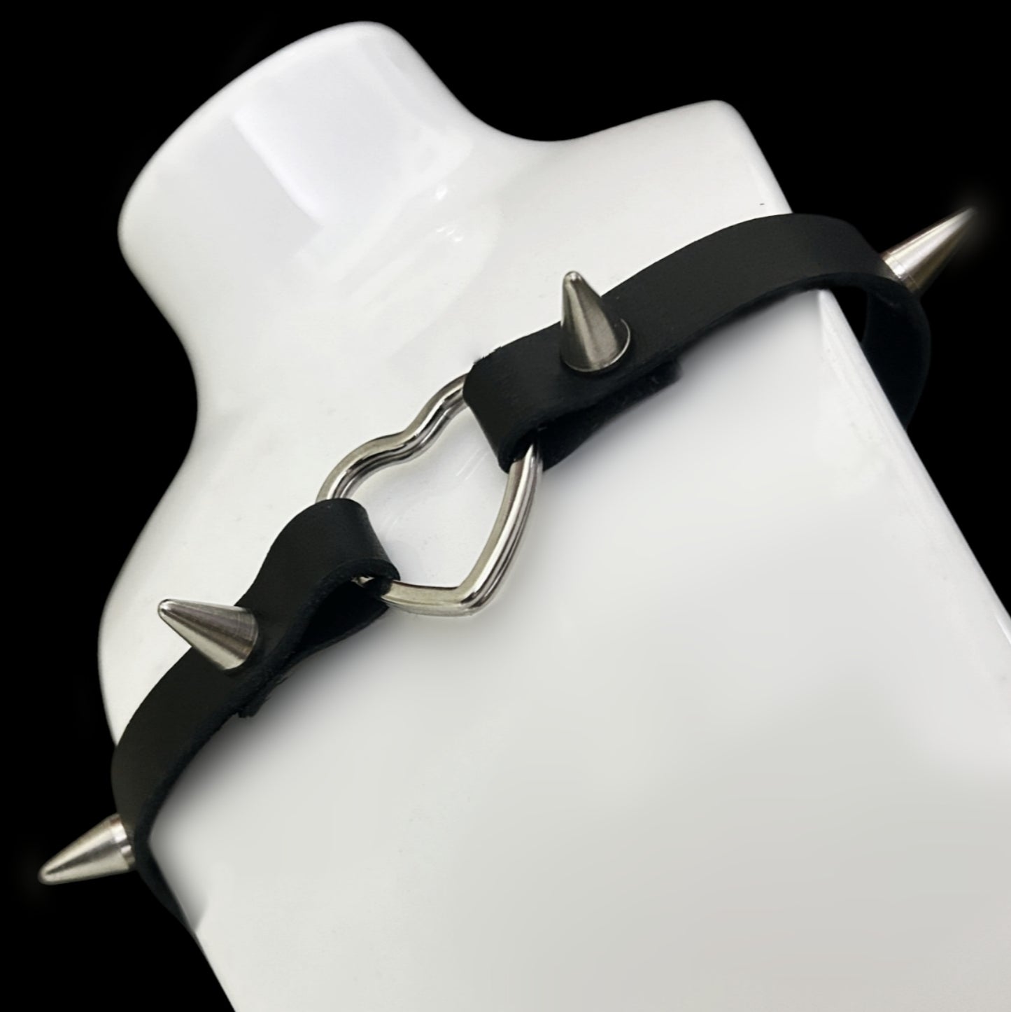 Choker displayed on a white bust, showing soft grain black leather strap with silver heart centerpiece and four spiked studs — a gothic, punk, and alt fashion statement for Y2K and cosplay aesthetics.
