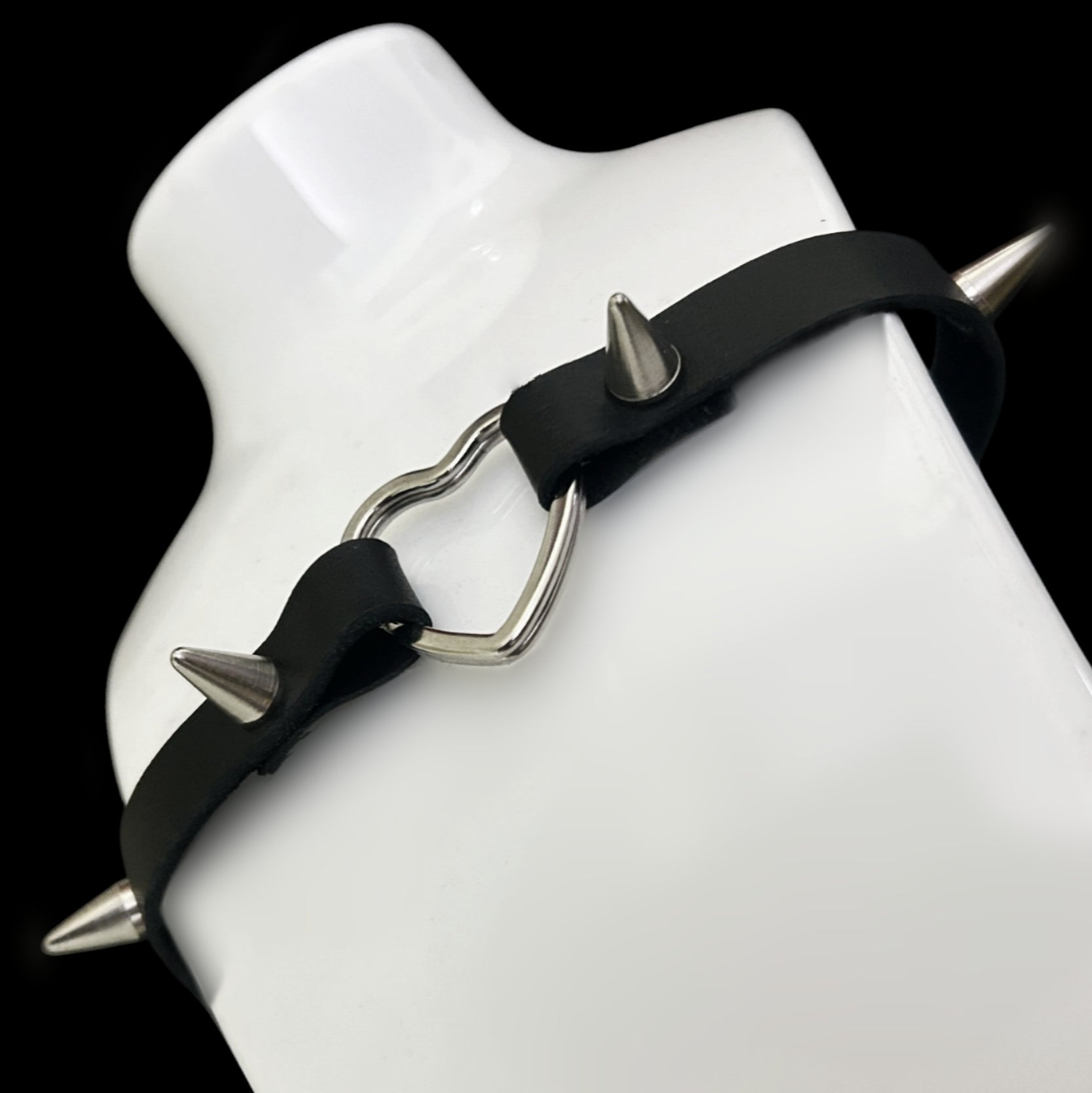 Choker displayed on a white bust, showing soft grain black leather strap with silver heart centerpiece and four spiked studs — a gothic, punk, and alt fashion statement for Y2K and cosplay aesthetics.