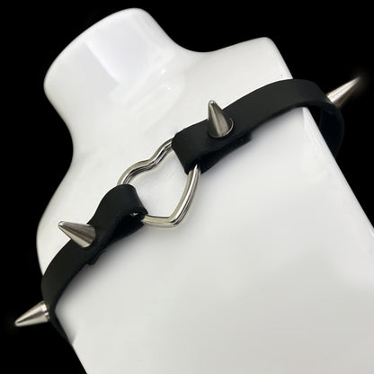 Choker displayed on a white bust, showing soft grain black leather strap with silver heart centerpiece and four spiked studs — a gothic, punk, and alt fashion statement for Y2K and cosplay aesthetics.